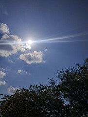 A bright sun shining in a clear blue sky with scattered clouds and tree silhouettes, natural daylight atmosphere