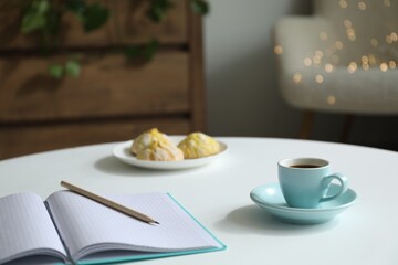 Notebook with pencil, cup of coffee and cookies on white table against blurred lights