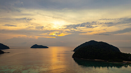 Fototapeta premium Aerial view of a stunning sunset above the Surin Islands. With golden hues reflecting on the calm ocean waters. The coral reef at the shoreline in the foreground adds depth. Paradise for snorkel