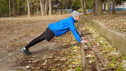 Man in sportswear performing incline push-ups on bench in autumn park for outdoor fitness training. Side view