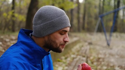 Close-up of man in grey beanie and blue jacket eating red apple in autumn park after outdoor workout. Side view portrait