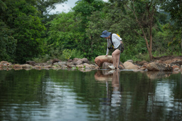 Young woman carrying a yellow backpack and camera is hiking through nature, carefully crossing a rocky stream in the forest. Outdoor adventure and eco-travel lifestyle concept.