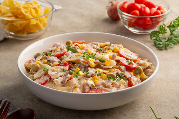 Homemade macaroni salad with bow tie pasta and canned tuna, carrot, onion, corn, tomato, green peas and mayonnaise dressing in a white plate on wooden table.
