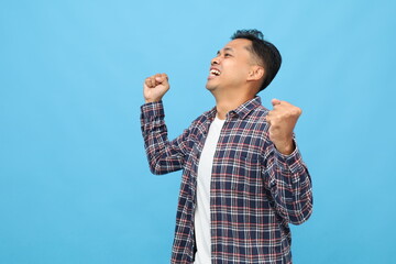 Side view of happy young Asian man with clenched fists raised up, shouting in celebration of success, standing on blue background.