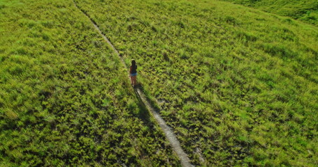 Aerial view capturing a female tourist walking along a winding path on a lush green hill, fully immersed in the breathtaking landscape on a bright, sunny summer day
