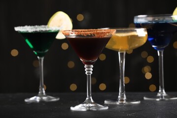 Different martini cocktails in glasses on black table against blurred lights, closeup