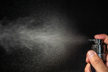 A close-up shot of a hand holding and spraying a perfume or liquid from a black bottle, with a fine mist visible against a dark background.

