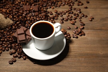 Aromatic coffee in cup, beans and pieces of chocolate on wooden table, closeup. Space for text