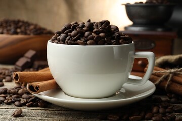 Aromatic coffee beans in cup, cinnamon and grinder on wooden table, closeup