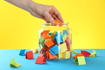 Woman taking orange paper piece from glass jar on color background, closeup
