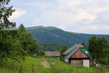 A rustic house with a gable roof and a barn, surrounded by green trees, stands on a hillside. Majestic mountains are visible in the background, with light clouds floating above them.