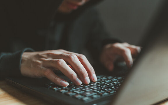 Hacker hands typing on laptop keyboard in dark environment. Cybercrime concept showing illegal computer activities, unauthorized access, data theft and malicious hacking operations.