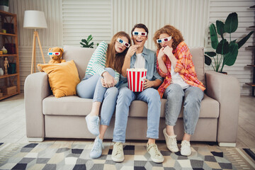 Naklejka premium Three friends enjoying a fun movie night at home wearing 3D glasses seated on a cozy sofa with popcorn and drinks
