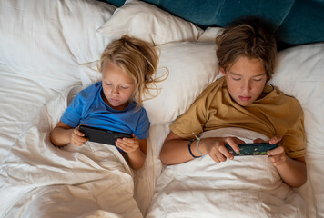 Two young children playing video games in bed under a cozy white blanket, captured from a top view, enjoying their leisure time together