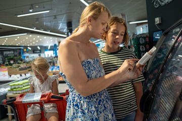 Mother and son carefully reading food label while choosing frozen product from supermarket refrigerator, daughter sitting in shopping cart