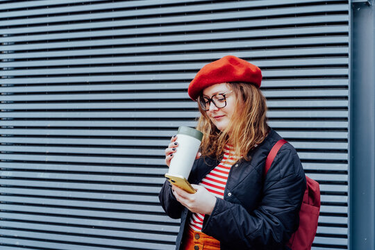 Young smiling plus size woman with backpack using mobile phone, drinking coffee outdoors on the street. Confident fashion hipster messaging, chatting, working remotely. Using navigator app for travel.