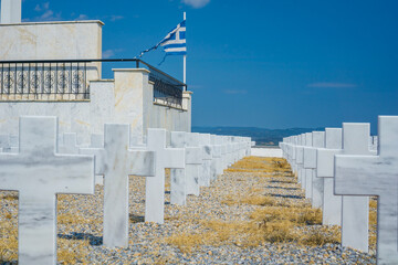 A row of white marble crosses at a greek cemetery under a bright blue sky, with a greek flag in the background, concept of business funeral services, religious articles, faith based organizations.