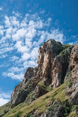 Naklejka premium Majestic rocky mountain peak with green slopes under blue sky and white scattered clouds