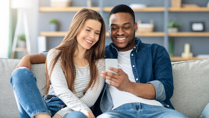 Portrait Of Happy Young Interracial Couple With Smartphone Relaxing At Home Together, Cheerful Multicultural Lovers Browsing Social Networks Or Using New App While Resting On Sofa In Living Room
