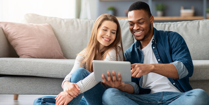 Happy Multiracial Couple Relaxing With Digital Tablet In Living Room, Cheerful Young Black Man And White Woman Browsing Social Networks On Tab Computer Or Shopping Online, Sitting On Floor At Home