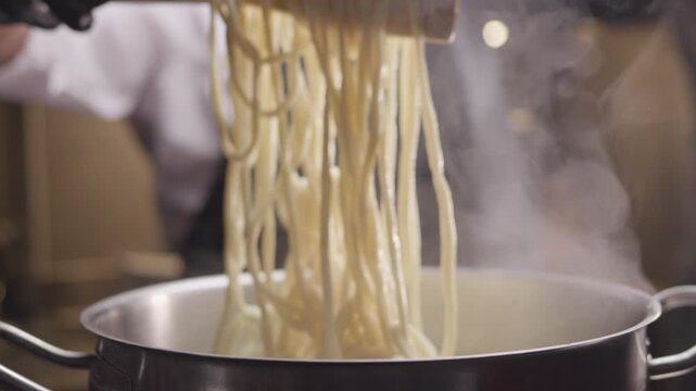 Close-up hands of chef in gloves and apron while stirring handmade pasta in the saucepan with boiling water. Restaurant of traditional Asian kitchen.