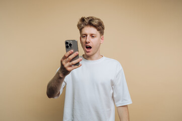 Young man expressing surprise while looking at his smartphone in a neutral-toned background during daytime