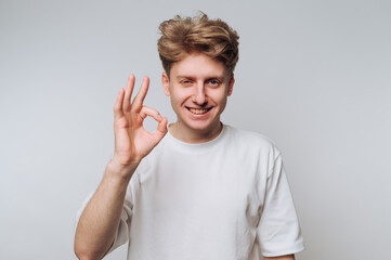 Smiling young man signals okay gesture against a neutral background during casual indoor setting