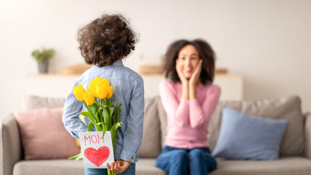 Surprise For Mommy. Rear view of African American daughter holding and hiding bouquet of yellow tulips flowers and greeting card behind her back, greeting exicted woman who sitting on the sofa