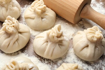 Uncooked khinkalis (dumplings) with rolling pin and flour on wooden table, closeup