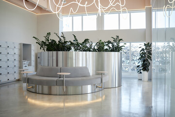 Modern waiting area featuring circular metallic bench with integrated tables and large planter filled with green plants, illuminated by abstract ceiling lights, empty of people