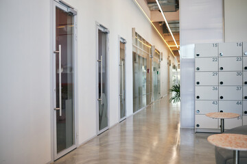Modern office hallway featuring glass doors along walls, polished concrete floor reflecting light, row of numbered lockers on right side, empty corridor extending into distance