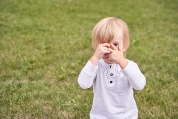 Fototapeta premium Blonde caucasian toddler playing peekaboo in park wearing white long sleeve shirt on a sunny day.