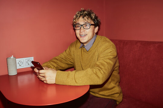 Portrait of young adult Asian man with curly hair and glasses sitting at table holding smartphone smiling looking toward camera in modern red interior