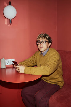 Portrait of young adult Asian man with curly hair and glasses sitting at table holding smartphone, looking toward camera, modern wall lamp and power outlets visible in background