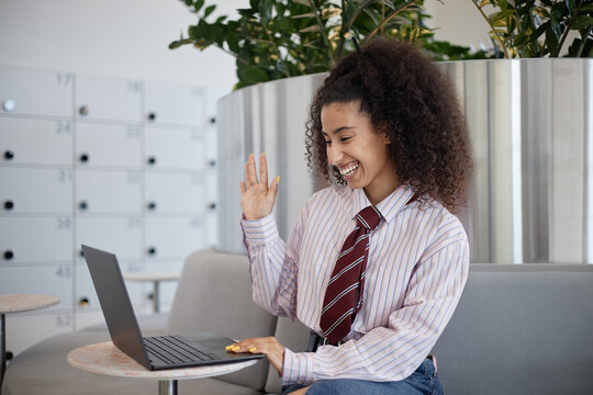 Young adult woman smiling and waving hand while sitting in modern office space, engaging in video call on laptop, curly hair visible, casual business attire, lockers in background