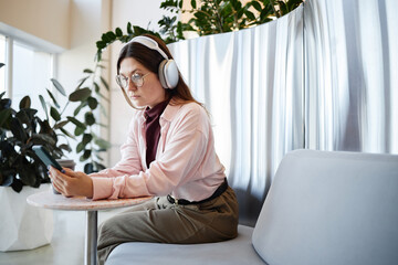 Young adult Caucasian woman wearing headphones sitting on sofa using digital tablet in modern indoor space, focusing on screen with serious expression, surrounded by green plants