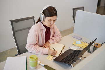 Caucasian young adult woman wearing headphones working at desk, writing in notebook while looking at laptop screen, surrounded by office supplies in modern workspace