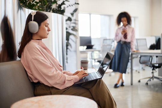 Caucasian young adult woman wearing headphones working on laptop while sitting on bench in modern office, biracial woman standing in background using smartphone near desks