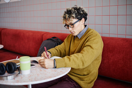 Young adult Asian man sitting at table writing in notebook, wearing glasses and concentrating on studying, backpack and headphones on side, reusable cup on table
