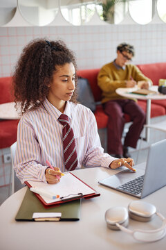 Young adult woman working on laptop and writing notes in notebook at table, sitting in modern workspace with young adult man studying in background, both focused on tasks