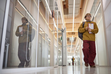 Young adult man walking down modern office hallway holding clipboard and backpack, looking forward with focused expression, reflection visible in glass wall