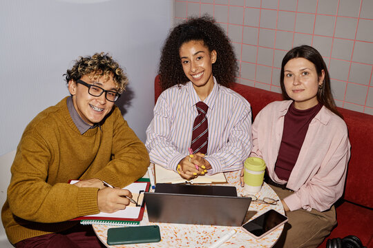 Three young adults, including Black woman and two Caucasian individuals, sitting at table collaborating on project, using laptop, notebooks, smartphone, smiling and looking at camera - Powered by Adobe