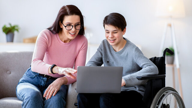 Home schooling concept. Disabled teen boy in wheelchair studying new online materials together with his mother indoors. Handicapped teenager and private tutor doing assignment on laptop