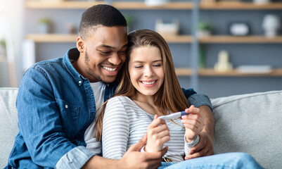 Happy multicultural couple holding positive pregnancy test and embracing on couch, romantic young black man and white woman rejoicing becoming parents, hugging while sitting on sofa at home, closeup