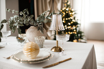 Kitchen table with dinner christmas set over xmas tree with glowing lights in room close up. Winter holiday season.