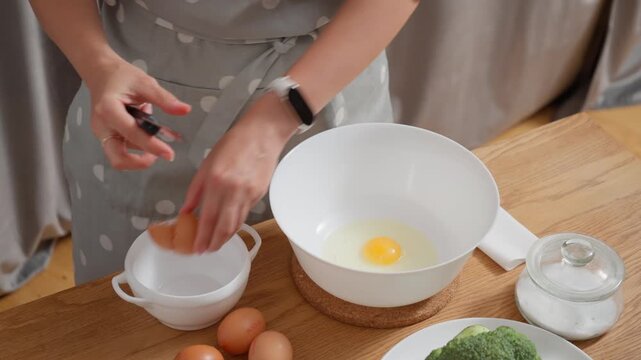 Female hands cracking raw eggs into a bowl. A housewife prepares ingredients for a quiche with broccoli, cooking a healthy breakfast or a homemade meal in the domestic kitchen.