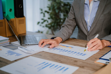 Focused Asian businesswoman reviews financial documents at desk, working through stacks of paperwork. Laptop open, pen in hand, analyzes reports, managing busy workload in modern office setting.