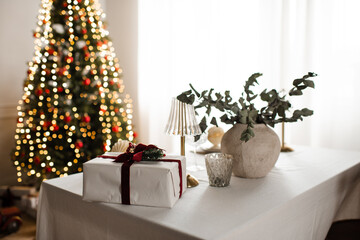 Christmas present box on table over xmas tree with glowing lights at home. Winter holiday season.