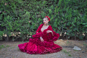Fototapeta premium Young, beautiful, blonde woman in red flamenco dress, flowers in her hair and gold jewelry, performing flamenco poses sitting on the ground with green plants in the background. Concept dance, art.