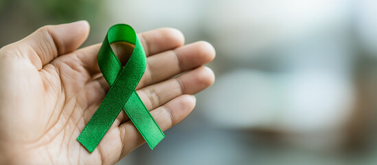 Hand holding green awareness ribbon against blurred background  